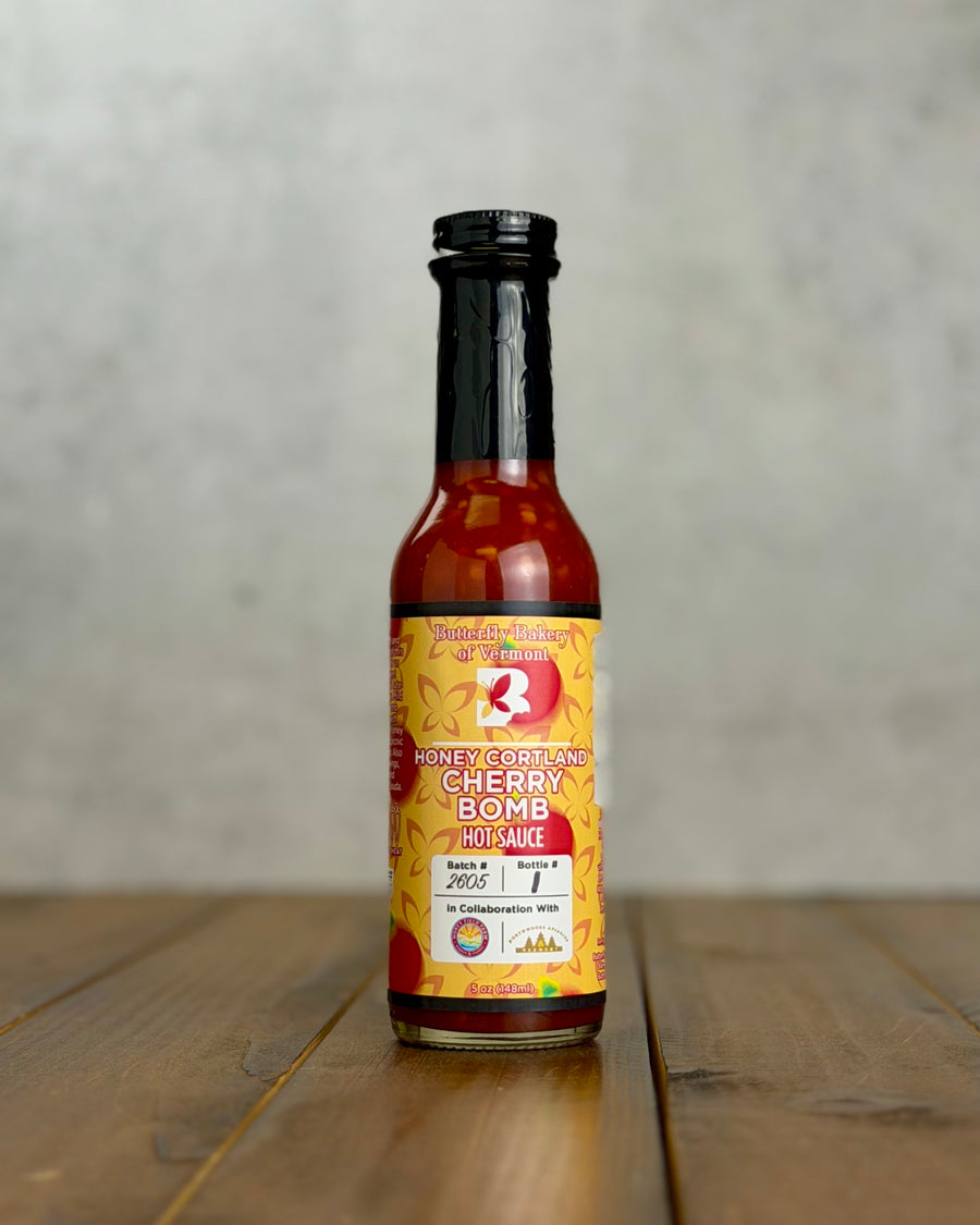 Bottle of Honey Cortland Cherry Bomb hot sauce from Butterfly Bakery of Vermont on a wooden tabletop
