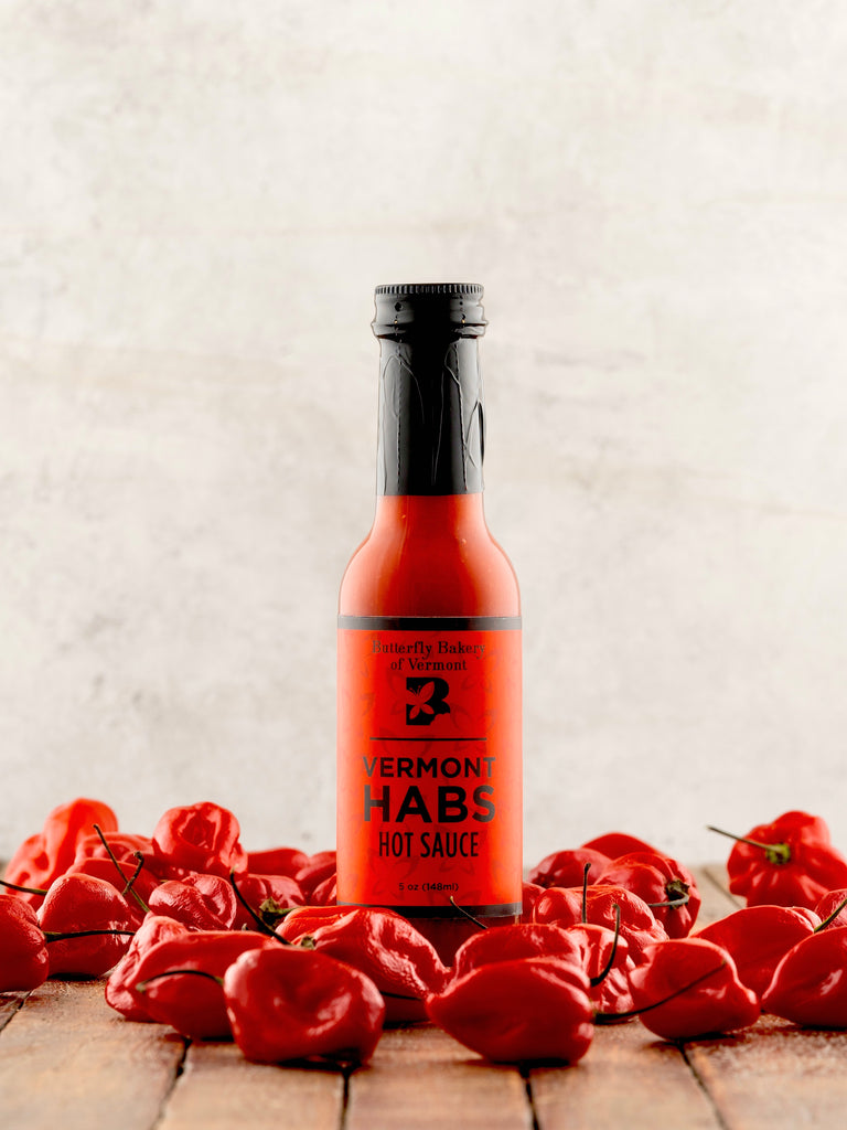 Bottle of Vermont Habs hot sauce surrounded by red peppers on a wooden surface.