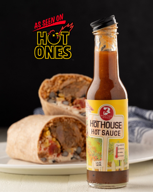 Bottle of Butterfly Bakery of Vermont Hot House hot sauce with a 'As Seen on Hot Ones' badge sitting on a table alongside a burrito cut in half on a plate