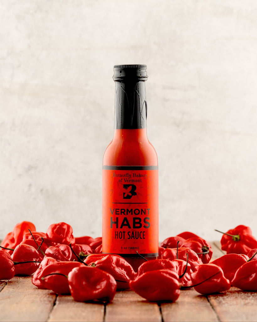 Bottle of Vermont Habs hot sauce surrounded by red peppers on a wooden surface.