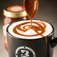 Fat Toad Farm caramel sauce being spooned into a foamy latte in a Butterfly Bakery of Vermont coffee mug