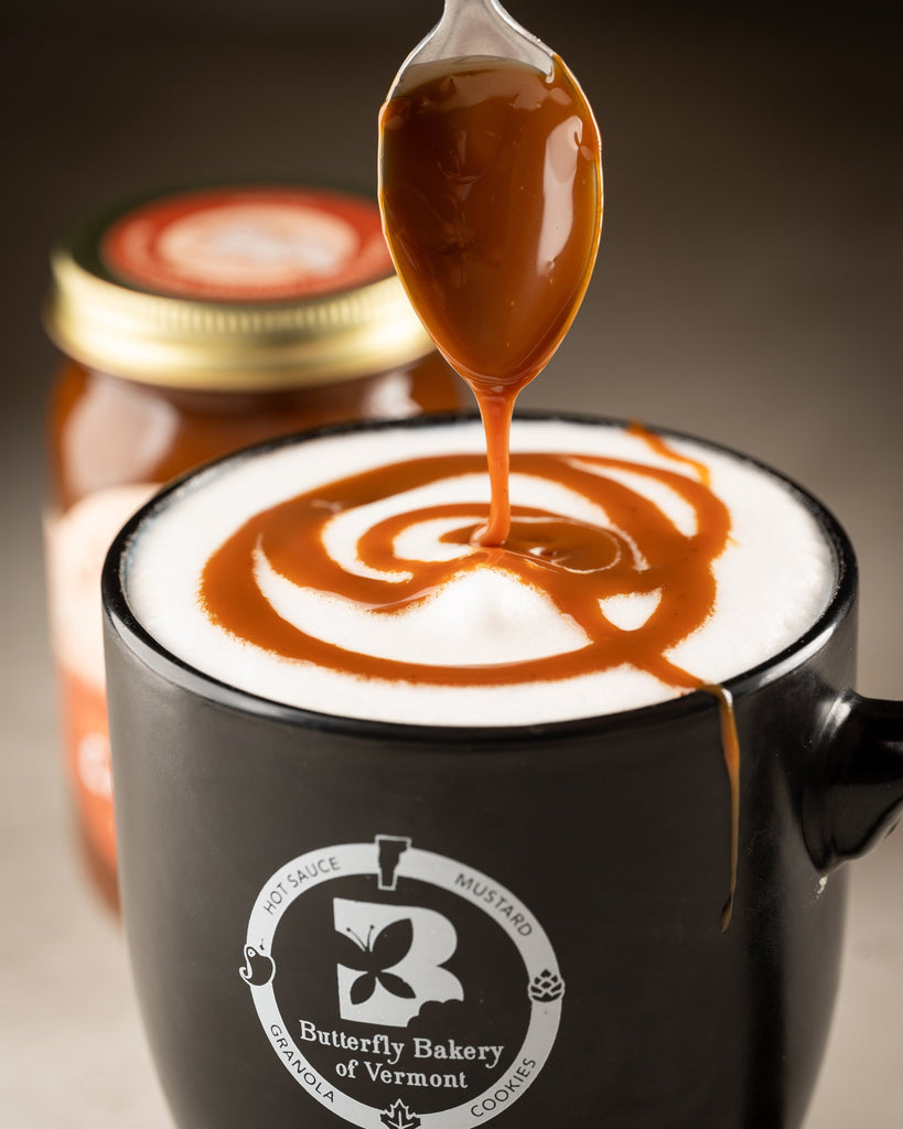 Fat Toad Farm caramel sauce being spooned into a foamy latte in a Butterfly Bakery of Vermont coffee mug