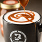 Fat Toad Farm caramel sauce being spooned into a foamy latte in a Butterfly Bakery of Vermont coffee mug