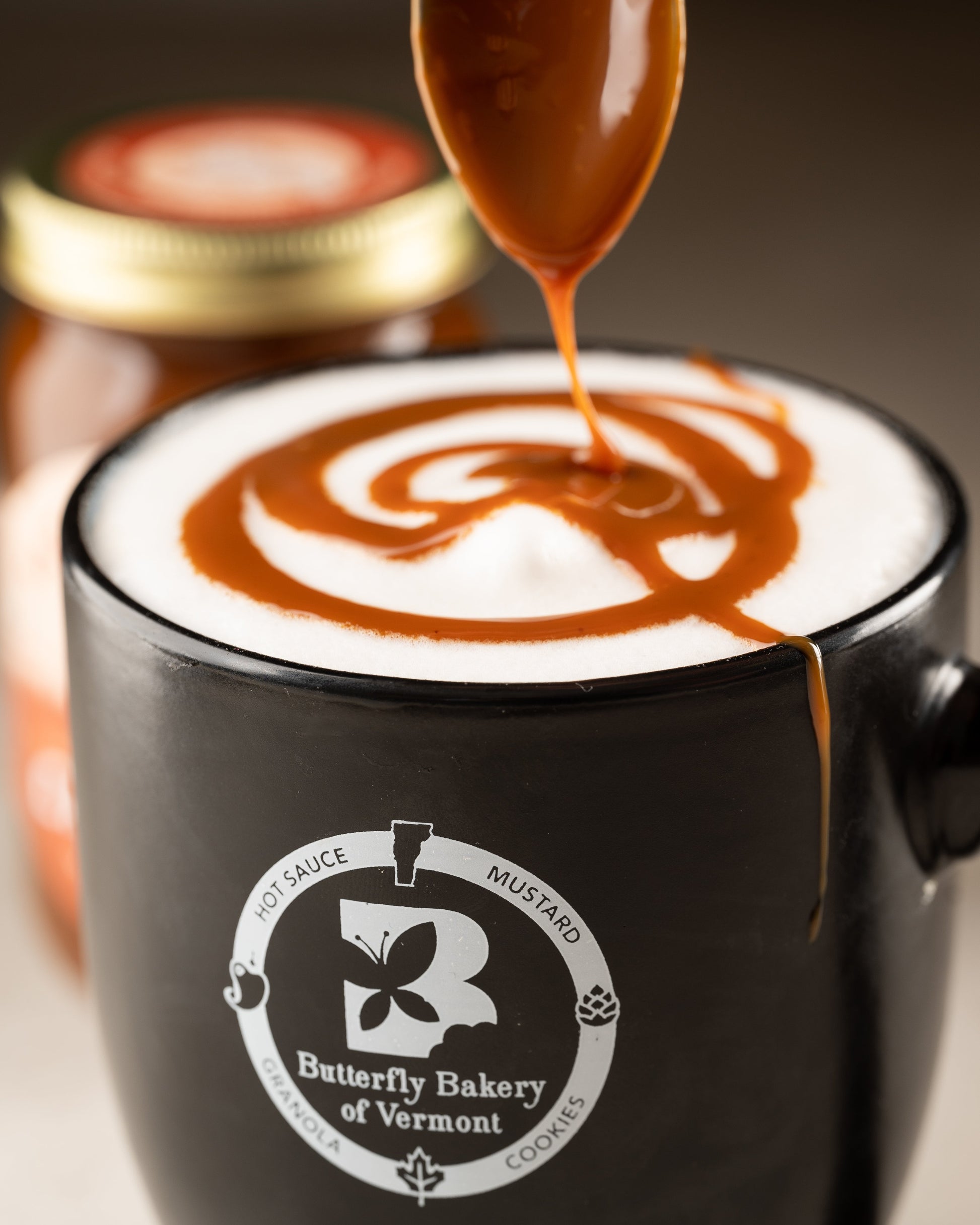 Fat Toad Farm caramel sauce being spooned into a foamy latte in a Butterfly Bakery of Vermont coffee mug