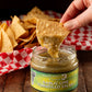 Tortilla chip being dipped into a jar of Butterfly Bakery of Vermont Garlic Lime Tomatillo Salsa sitting on a wood table along side a basket of tortilla chips