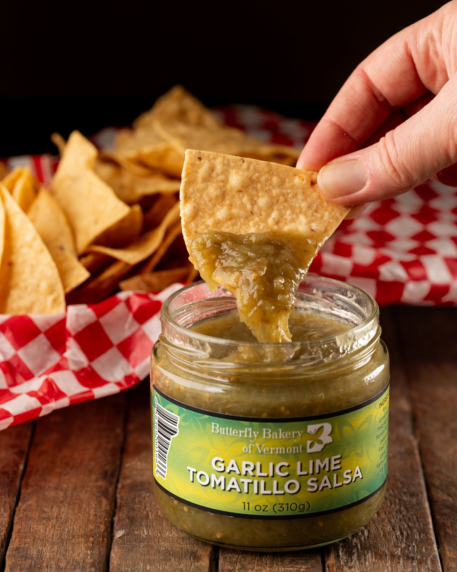 Tortilla chip being dipped into a jar of Butterfly Bakery of Vermont Garlic Lime Tomatillo Salsa sitting on a wood table along side a basket of tortilla chips