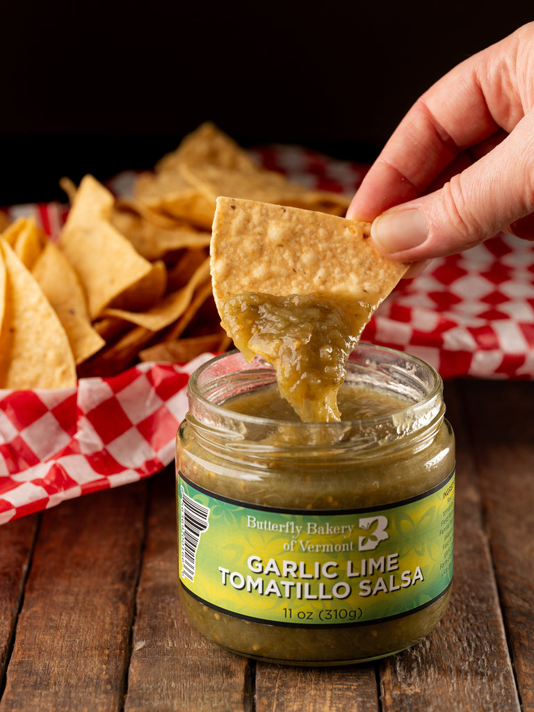 Tortilla chip being dipped into a jar of Butterfly Bakery of Vermont Garlic Lime Tomatillo Salsa sitting on a wood table along side a basket of tortilla chips
