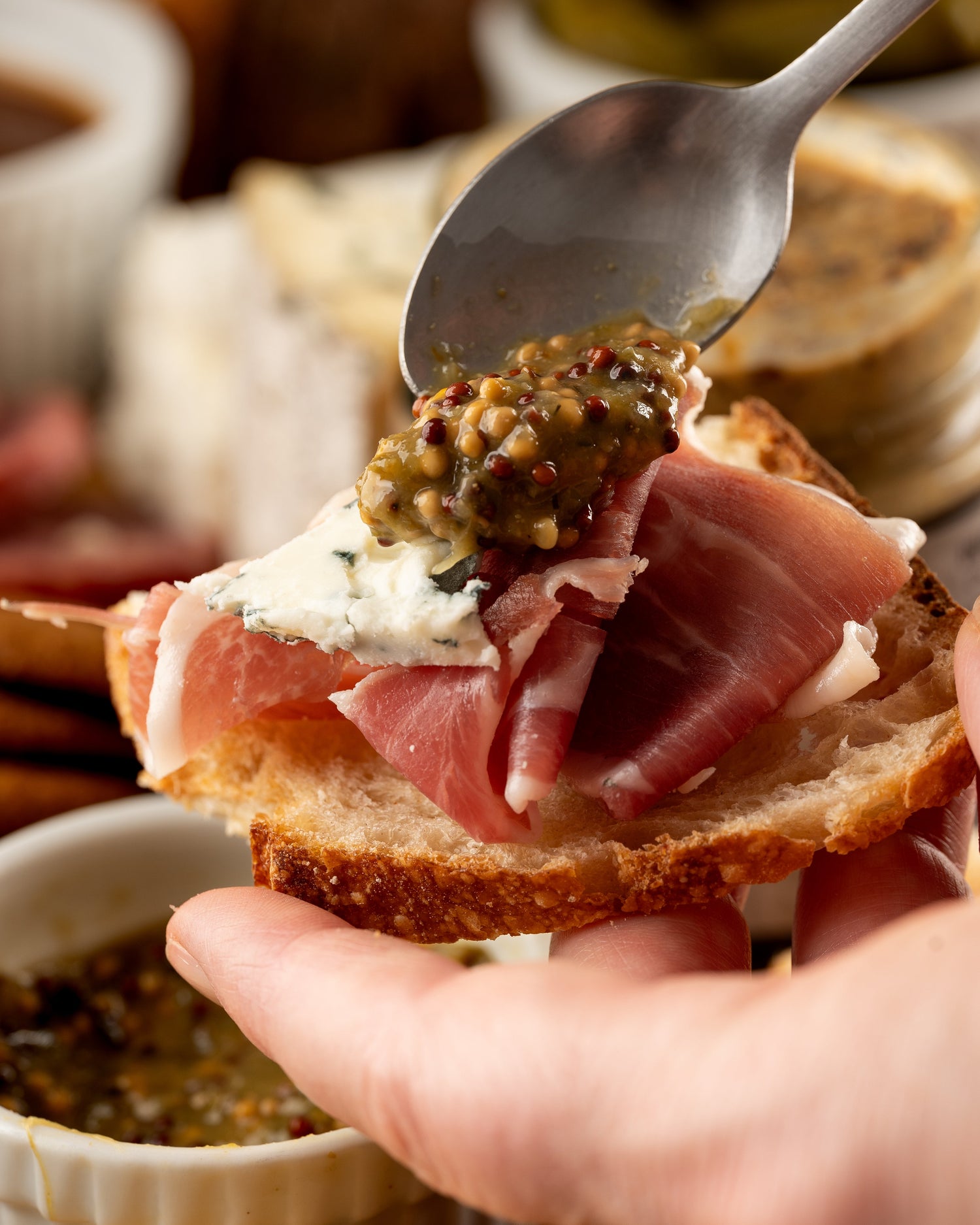 Butterfly Bakery of Vermont whole grain mustard being spooned onto brie and prosciutto on a slice of rustic bread