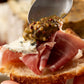 Butterfly Bakery of Vermont whole grain mustard being spooned onto brie and prosciutto on a slice of rustic bread