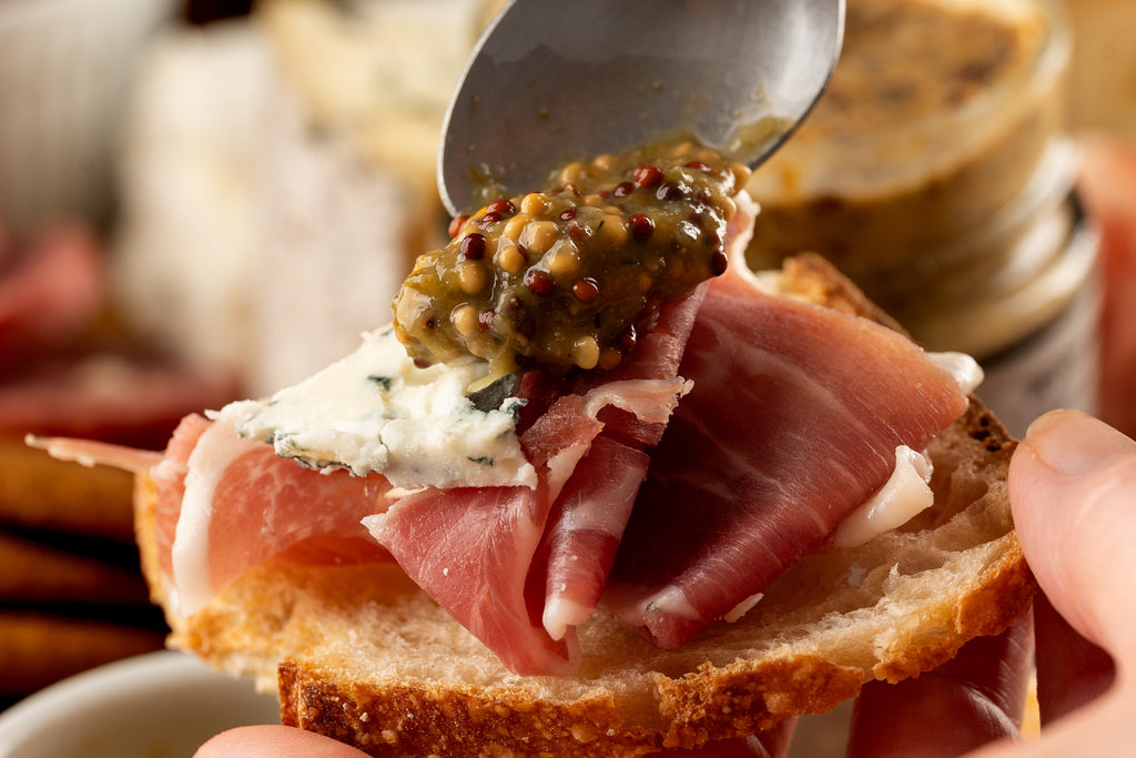 Butterfly Bakery of Vermont whole grain mustard being spooned onto brie and prosciutto on a slice of rustic bread