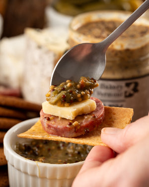 Hand holding a cracker with salami, cheese, and mustard being spooned ontop.