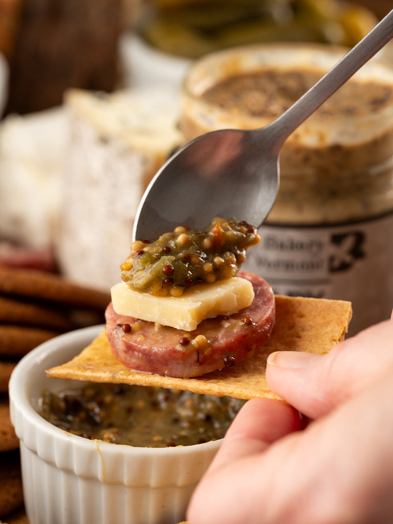 Hand holding a cracker with salami, cheese, and mustard being spooned ontop.