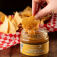 Tortilla chip being dipped into a jar of Butterfly Bakery of Vermont Tomatillo Chipotle Salsa sitting on a wood table along side a basket of tortilla chips