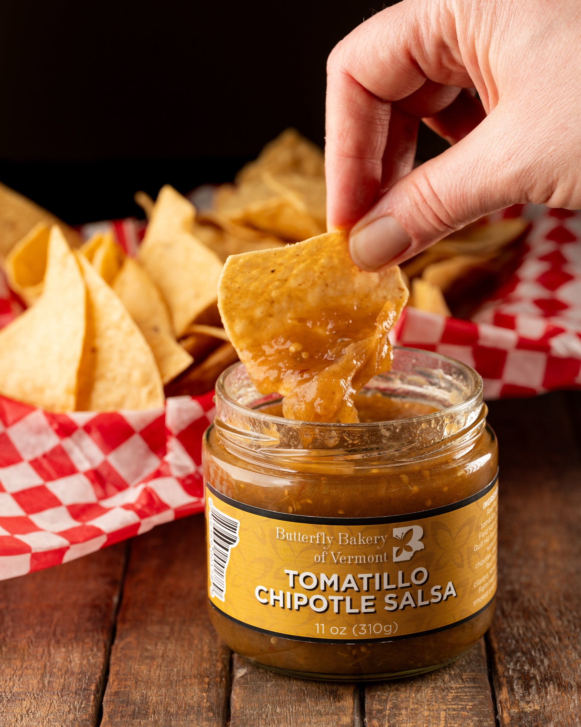 Tortilla chip being dipped into a jar of Butterfly Bakery of Vermont Tomatillo Chipotle Salsa sitting on a wood table along side a basket of tortilla chips