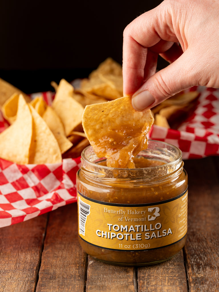 Tortilla chip being dipped into a jar of Butterfly Bakery of Vermont Tomatillo Chipotle Salsa sitting on a wood table along side a basket of tortilla chips