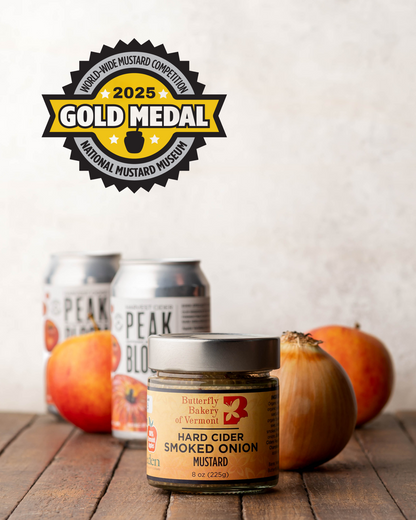 Jar of Butterfly Bakery of Vermont Hard Cider Smoked Onion Mustard with a gold metal badge sitting on a wood surface with 2 cans of hard cider, an onion and 2 apples