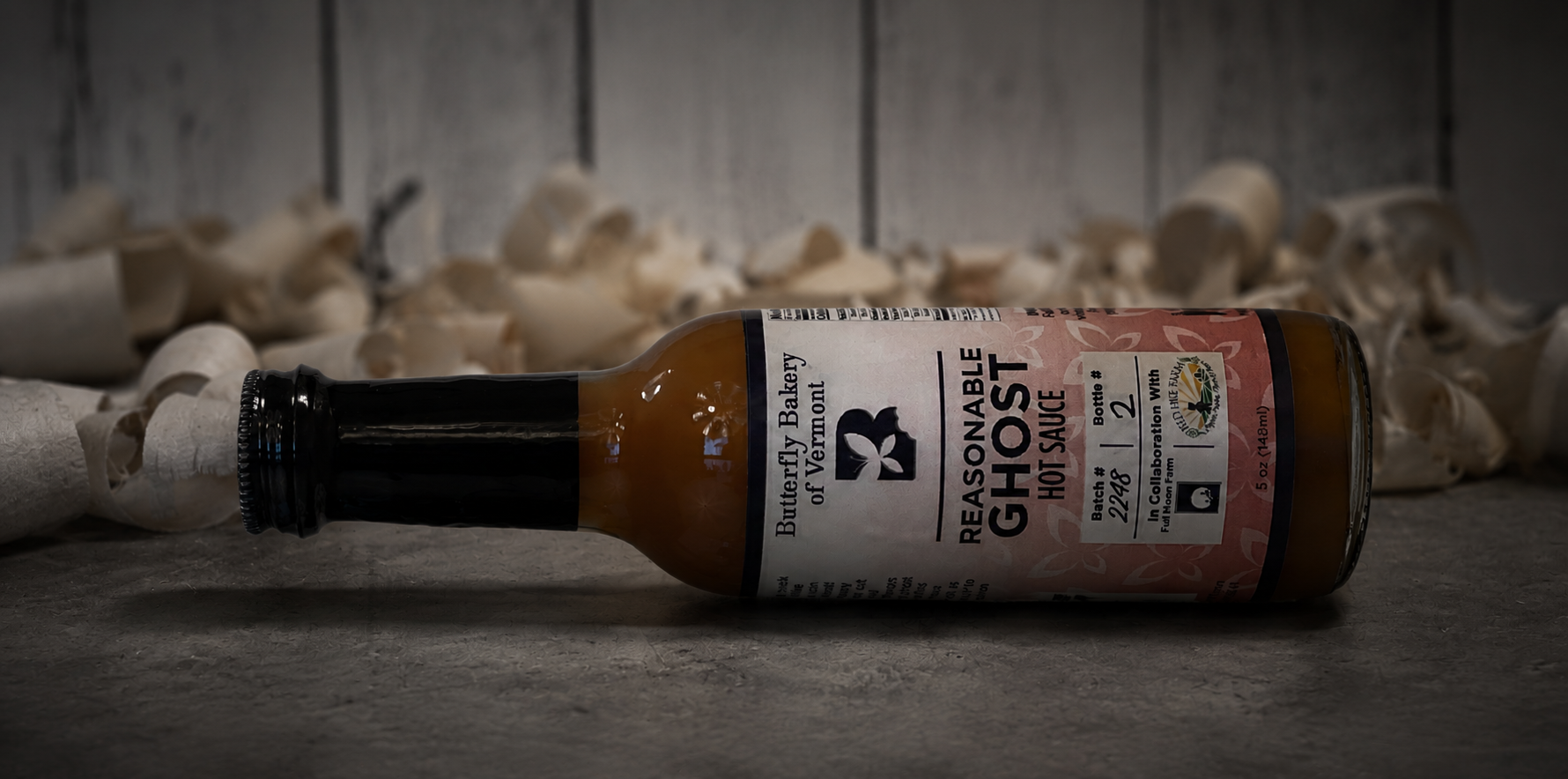A bottle of Reasonable Ghost Hot Sauce lies on its side on a dark tabletop, with blurred white mushrooms in the background against a rustic gray wood wall.