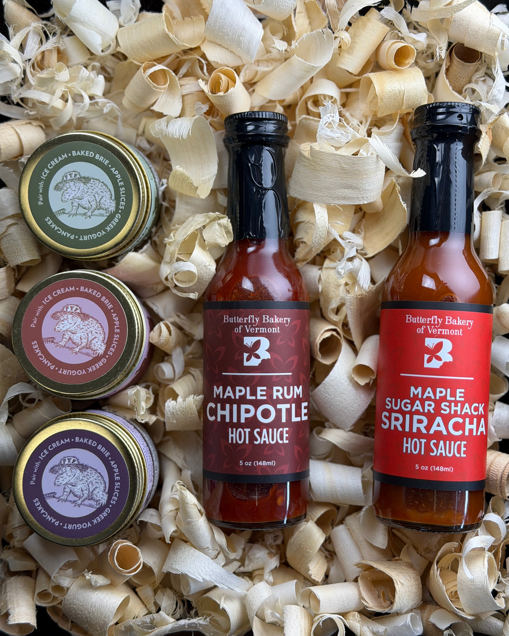 Two bottles of hot sauce and three small containers of caramel on a bed of wood shavings.